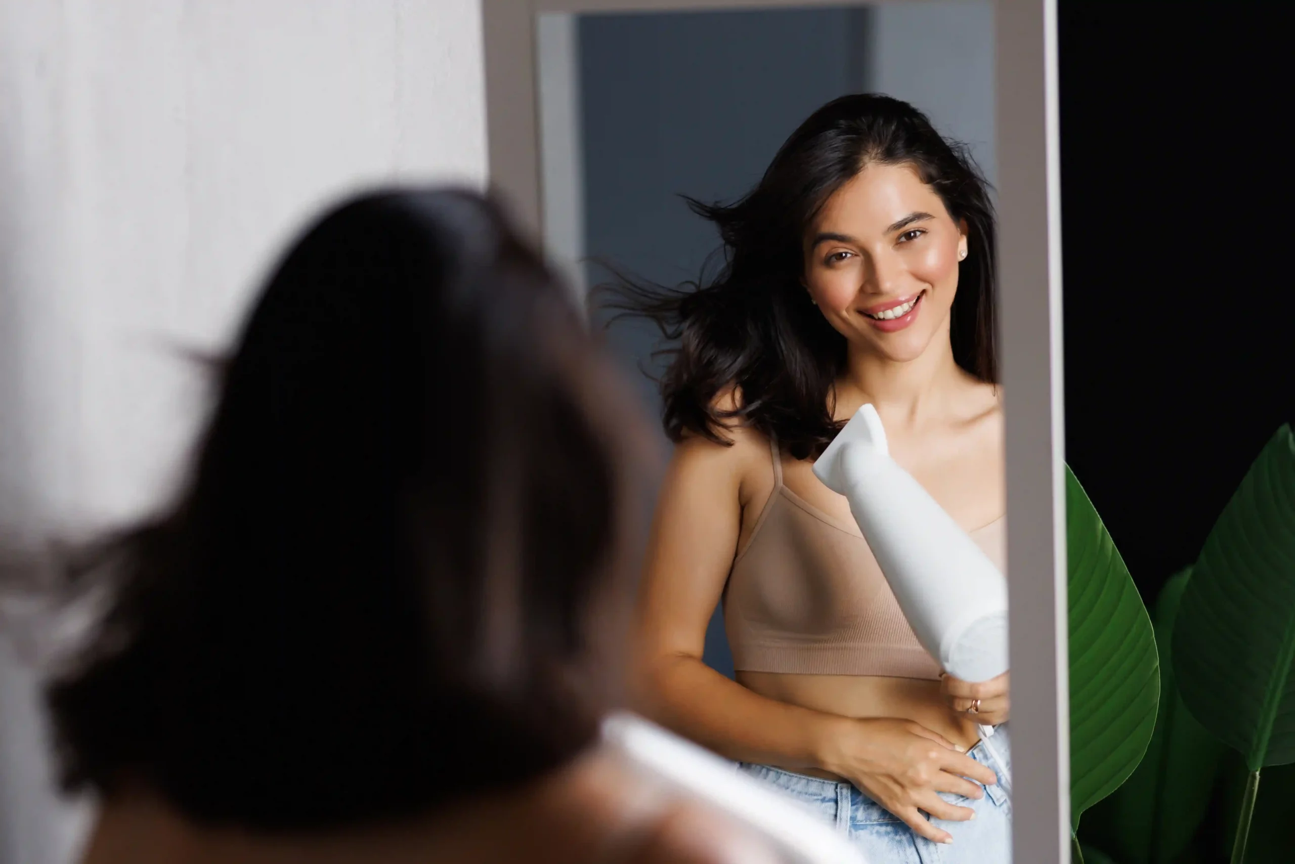 Woman smiling in mirror while styling hair with blow dryer